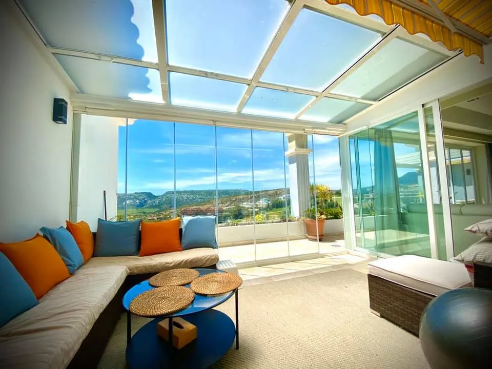 Sunlit interior beneath a glass roof, opening onto a terrace with hillside views